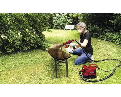 Frau verwendet ein Farbsprühsystem von Einhell, um eine Holzbank im Garten zu lackieren.