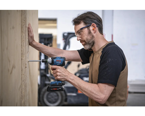 Ein Mann schraubt mit einem Akku-Schlagschrauber eine Schraube in ein Holzbrett. Er trägt dabei eine Schutzbrille und Ohrstöpsel. Bosch Logo.