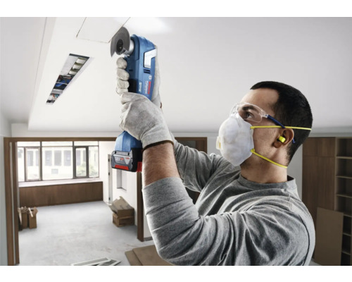 Mann schneidet mit einem Multifunktionswerkzeug eine Deckenöffnung, während er Sicherheitsausrüstung trägt.