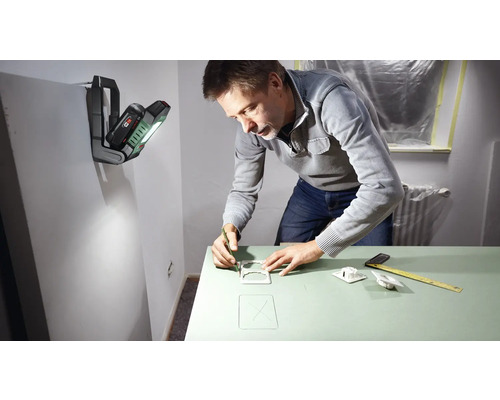 Ein Mann zeichnet mit Bleistift Umrisse auf eine grüne Oberfläche, beleuchtet von einer tragbaren Arbeitsleuchte an der Wand.