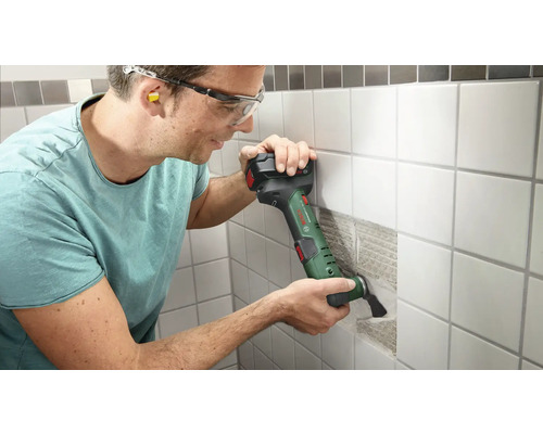 Mann arbeitet mit einem Multifunktionswerkzeug an einer gefliesten Wand. Er trägt dabei eine Schutzbrille und Ohrstöpsel.