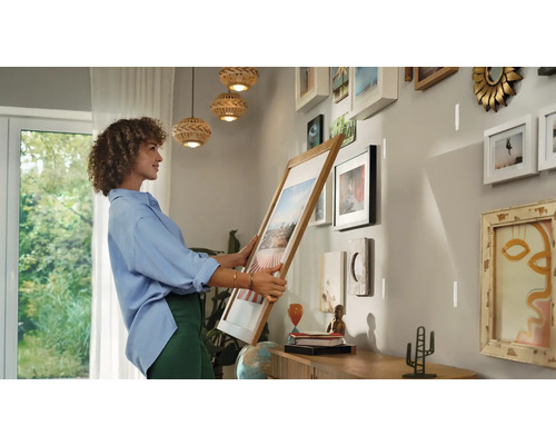 Frau hängt einen Bilderrahmen an einer Wand mit bereits befestigten Bildern auf.