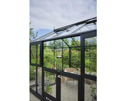 Gewächshaus mit Glaswänden und schwarzem Rahmen, ausgestattet mit Pflanzen und Sitzgelegenheiten.