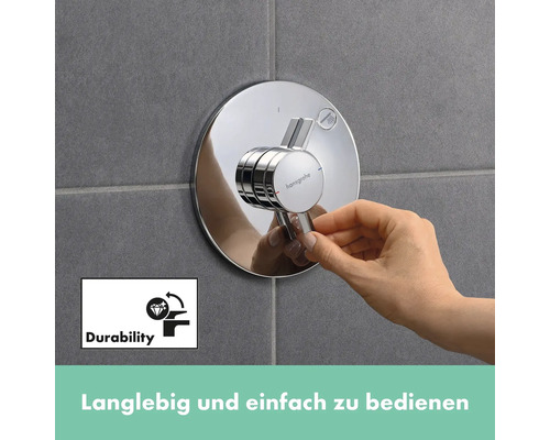 Nahaufnahme einer Hand, die einen runden, verchromten Hansgrohe Duschmischer an einer gefliesten Wand bedient. Das Symbol für Langlebigkeit ist ebenfalls im Bild zu sehen.