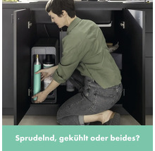 Eine Frau kniet vor einem geöffneten Küchenschrank und setzt eine CO2-Flasche in einen Wassersprudler ein.