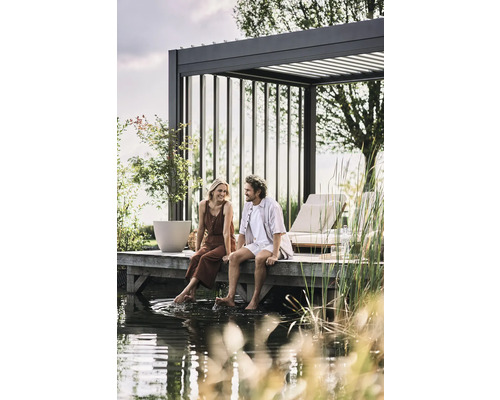 Paar sitzt auf einem Holzsteg am Wasser unter einer Pergola. Gartengestaltung mit Loungemöbeln und Bepflanzung.