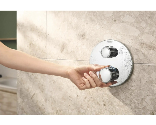 Eine Hand bedient einen runden, verchromten Duschthermostat von Grohe an einer gefliesten Wand.