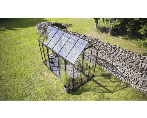 Gewächshaus auf einer Wiese mit einem Holzstapel im Hintergrund.