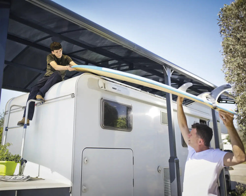 Zwei Personen hieven ein Surfbrett auf ein Wohnmobil mit Markise.