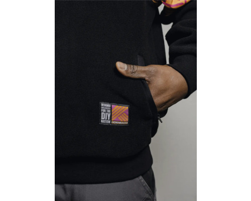Detailansicht einer schwarzen Fleecejacke mit Hornbach DIY Nation Aufnäher und einer Hand in der Tasche