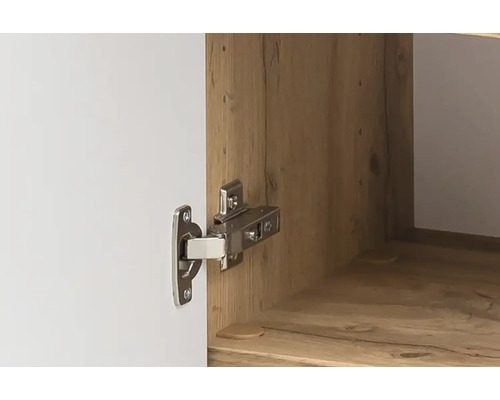Nahaufnahme eines Topfscharniers aus Metall an einer weißen Schranktür und einem Korpus in Holzoptik.
