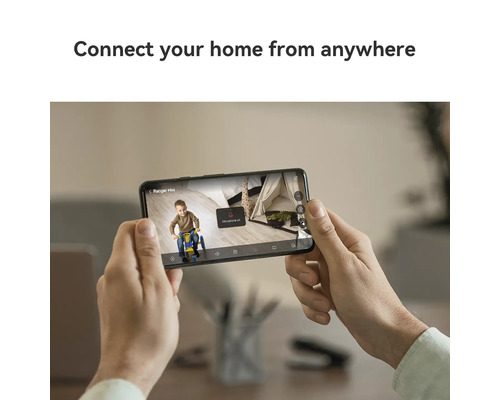 Eine Person hält ein Smartphone mit der Anzeige eines Kindes auf einem Dreirad vor einem Zelt.
