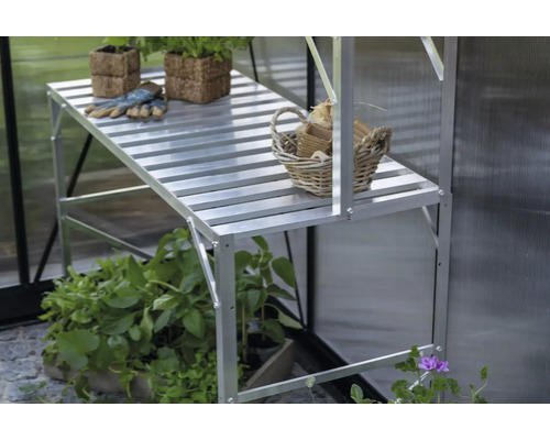 Aluminiumregal im Gewächshaus mit Pflanzen in Fasertöpfen, Gartenhandschuhen und einem geflochtenen Korb auf der Ablagefläche.