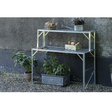 Zweistufiger Pflanztisch aus Aluminium im Freien vor einer Wand mit Gartenutensilien und Pflanzen in Töpfen auf Kiesboden.