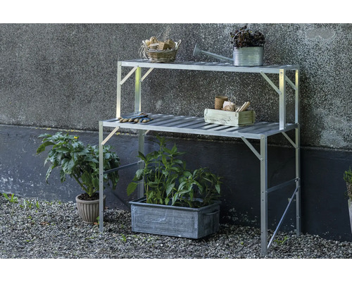 Zweistufiger Pflanztisch aus Aluminium im Freien vor einer Wand mit Gartenutensilien und Pflanzen in Töpfen auf Kiesboden.