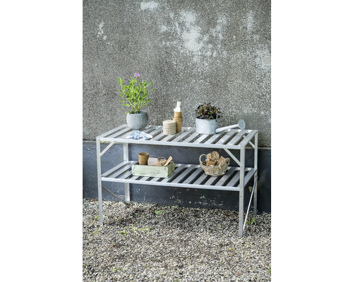 Zweistöckiges Gewächshausregal aus Aluminium mit Pflanzen, Gießkanne und Gartenutensilien auf einer Kiesfläche.