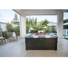 Moderner quadratischer Whirlpool mit zwei Personen auf einer hell gefliesten Terrasse inmitten von Gartenpflanzen.