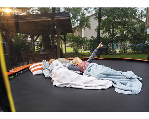 Kinder liegen auf einem großen Gartentrampolin mit Kissen und Decken im Garten neben einem Holzpavillon. Ein Kind zeigt in den Himmel.