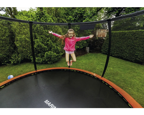Ein Kind springt auf einem runden Gartentrampolin mit Sicherheitsnetz und orangefarbenem Randschutz in einem grünen Garten.