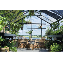 Helles Gewächshaus aus Glas und Metall mit zwei Korbsesseln, einem Holztisch und zahlreichen Pflanzen vor einer Aussicht auf einen See.