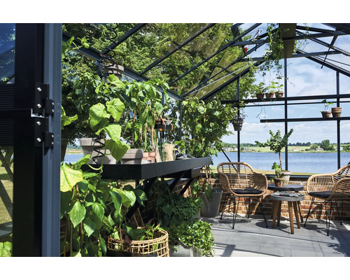 Helles Gewächshaus mit Glaswänden und Metallrahmen, bepflanzt mit diversen Pflanzen sowie Sitzbereich mit Rattansesseln und Blick auf einen See.