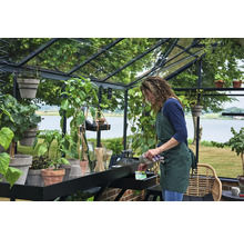 Frau mit Schürze bei der Gartenarbeit in einem hellen, organisierten Gewächshaus mit schwarzem Metallrahmen und verschiedenen Topfpflanzen.