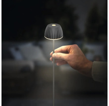 Hand hält eine moderne LED-Akkuleuchte mit geripptem Schirm in Anthrazit, die im Dunkeln leuchtet.
