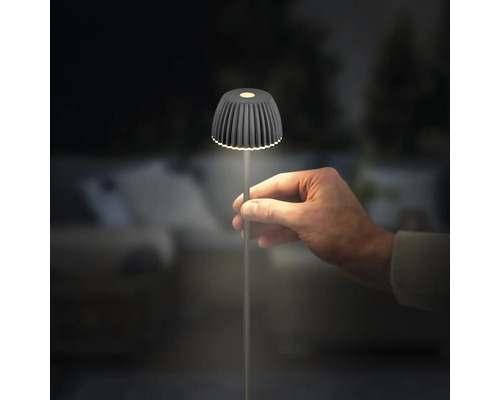 Hand hält eine moderne LED-Akkuleuchte mit geripptem Schirm in Anthrazit, die im Dunkeln leuchtet.