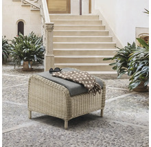 Geflochtener Gartenhocker mit grauem Kissen in einem hellen Innenhof mit Steintreppe und Pflanzen.