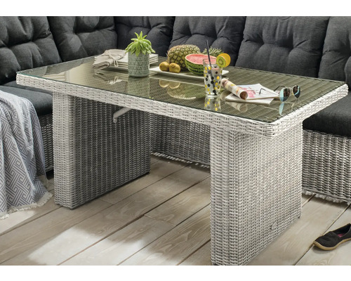 Gartentisch aus Polyrattan mit Glasplatte vor einer Lounge-Ecke mit dunklen Polstern auf einer Terrasse.