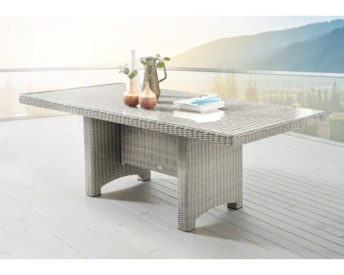 Rechteckiger Gartentisch aus Polyrattan-Geflecht mit Glasplatte auf einer hellen Terrasse vor einer Gebirgskulisse.