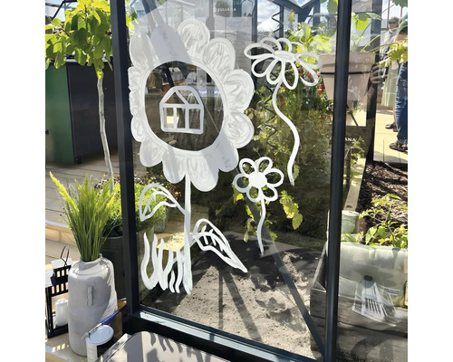 Mit weißer Farbe auf die Glasscheibe eines Gewächshauses gemalte stilisierte Blumen und ein kleines Haus, im Hintergrund Pflanzen.