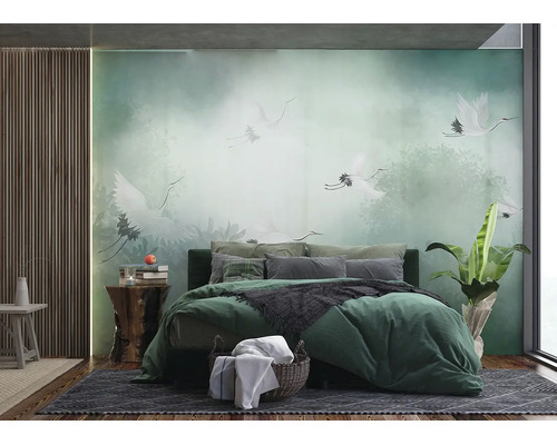 Fototapete im Schlafzimmer mit weißen Kranichen im nebligen Wald, grünes Bett mit Kissen, Holzpaneele an der Wand und Zimmerpflanze.