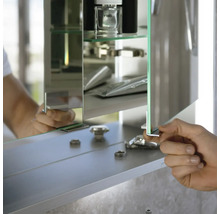 Badezimmer-Spiegelschrank mit Glasfachböden, eine Hand öffnet die verspiegelte Tür, Innenansicht mit Abstellfläche für Badutensilien.