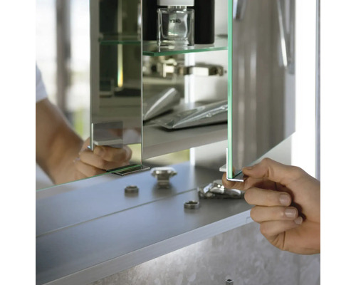 Badezimmer-Spiegelschrank mit Glasfachböden, eine Hand öffnet die verspiegelte Tür, Innenansicht mit Abstellfläche für Badutensilien.