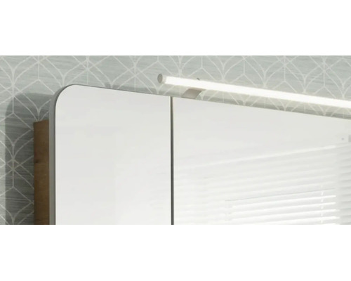 Spiegelschrank mit LED-Aufsatzleuchte und abgerundeten Ecken vor einer gemusterten Wand.