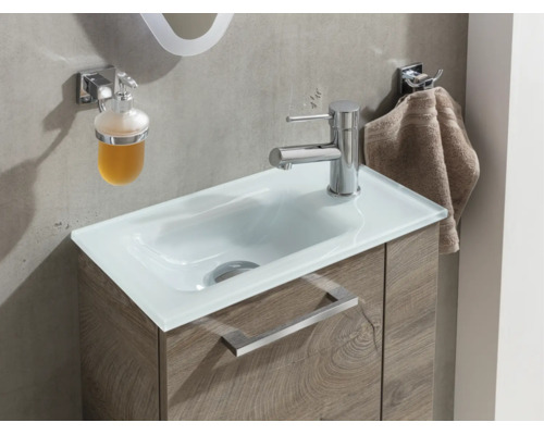 Moderner Waschtisch mit weißem Glasbecken, Unterschrank in Holzoptik, verchromter Armatur, Seifenspender und Handtuchhalter.