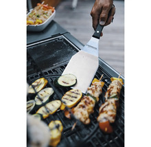 Hand führt Edelstahl Grillwender mit Campingaz Logo über Grillgemüse und Fleischspieße auf einem schwarzen Grillrost.