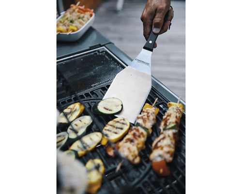 Hand führt Edelstahl Grillwender mit Campingaz Logo über Grillgemüse und Fleischspieße auf einem schwarzen Grillrost.