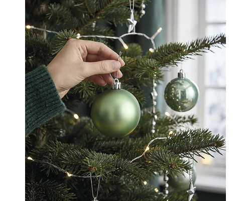 Eine Hand hängt eine matte grüne Weihnachtskugel an einen geschmückten Weihnachtsbaum mit LED Lichterkette.