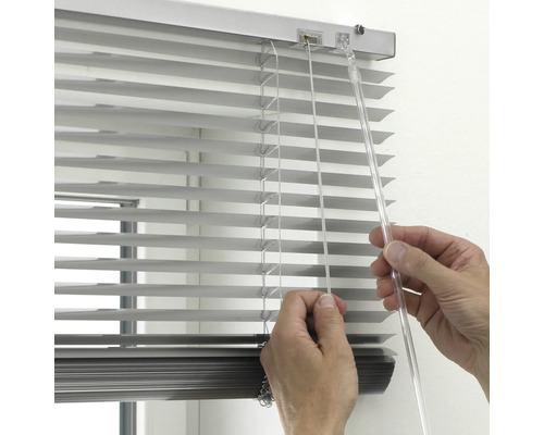 Nahaufnahme einer Hand, die eine silberne Aluminium-Jalousie mit transparentem Wendestab und Zugschnur bedient.