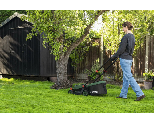 Frau mäht den Rasen mit einem Bosch Rasenmäher in einem Garten mit Holzschuppen und Baum.