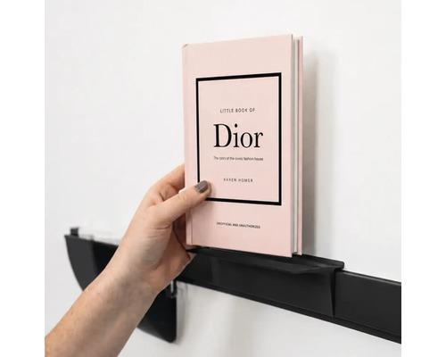 Eine Hand platziert ein Buch auf einem minimalistischen, schwarzen Wandregal aus Metall an einer Wand.