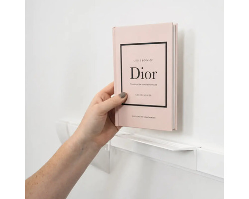 Eine Hand platziert das Buch Little Book of Dior auf einem schmalen weißen Wandregal.