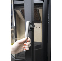 Hand an einem silbernen Stangengriff einer dunklen Kaminofentür mit Schließmechanismus.