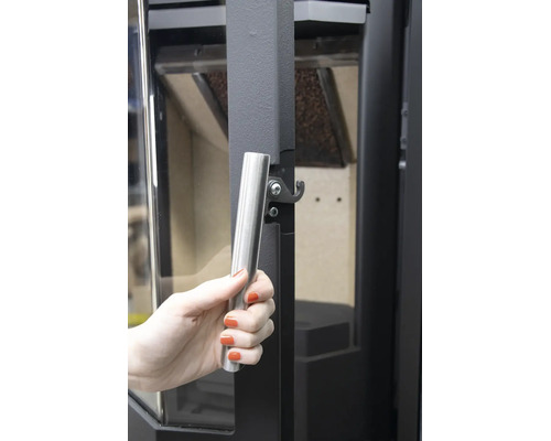 Hand an einem silbernen Stangengriff einer dunklen Kaminofentür mit Schließmechanismus.