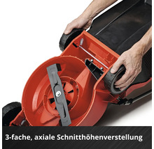 Unterseite eines Rasenmähers mit sichtbarem Schnittmesser und Händen, die die axiale Schnitthöhenverstellung am Rad demonstrieren.