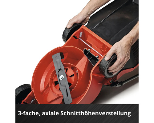 Unterseite eines Rasenmähers mit sichtbarem Schnittmesser und Händen, die die axiale Schnitthöhenverstellung am Rad demonstrieren.