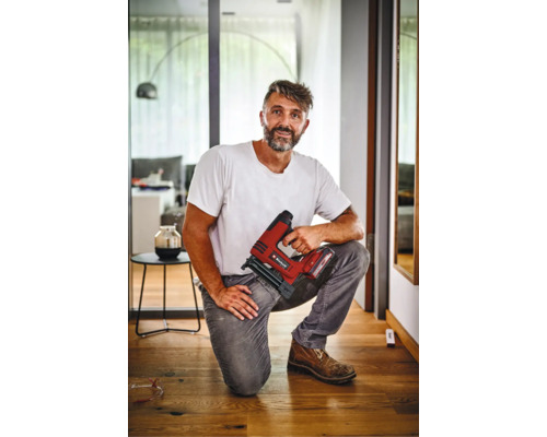 Handwerker kniet auf einem Holzboden und hält einen Akku Tacker der Marke Einhell in der Hand in einem modernen Wohnraum.