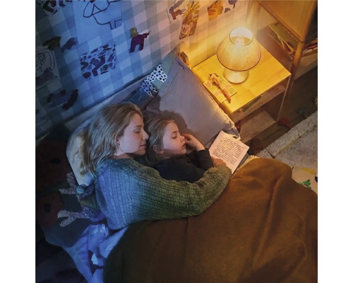 Mutter und Kind liegen im Bett und lesen bei gedimmtem Licht einer Nachttischlampe in einem gemütlichen Kinderzimmer.